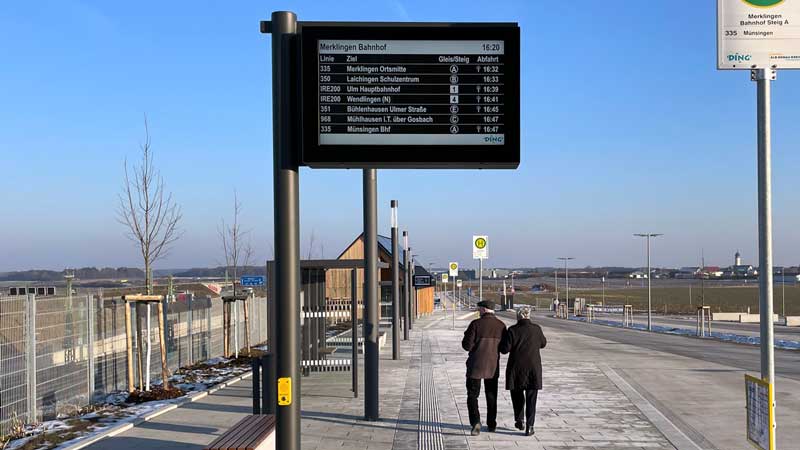 Anzeigetafel mit Busverbindungen, darunter läuft ein älteres Paar an der Bushaltestelle entlang